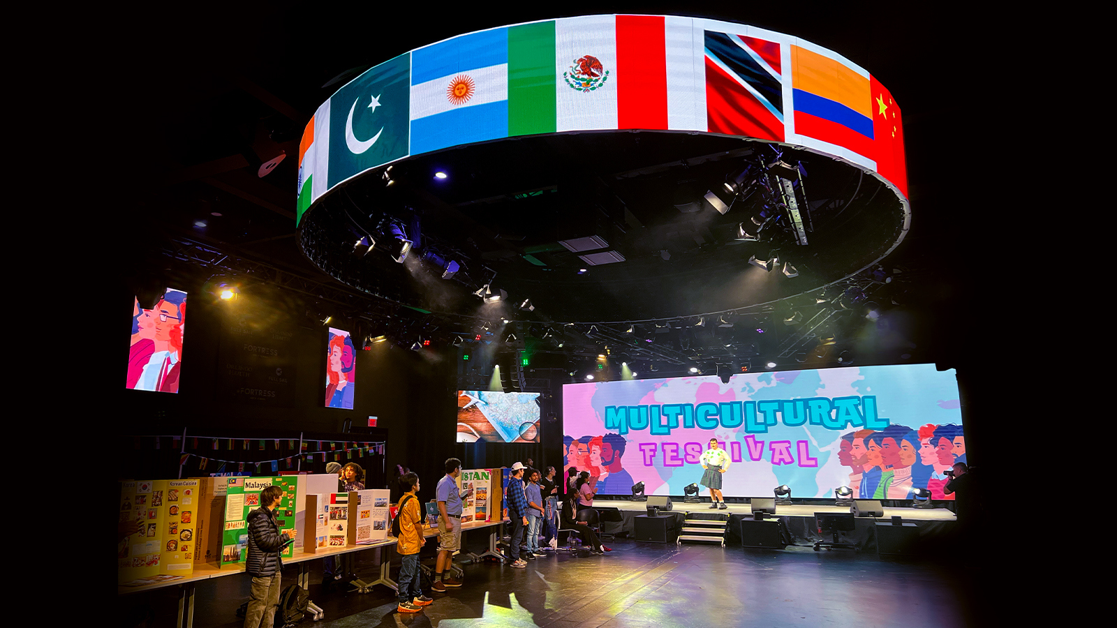 Students stand by a line of tables against a wall with cardboard presentations on various countries. One student poses onstage in a kilt and white shirt with a large screen behind them displaying “Multicultural Festival.” Above the room, a circular screen displays flags of various countries.