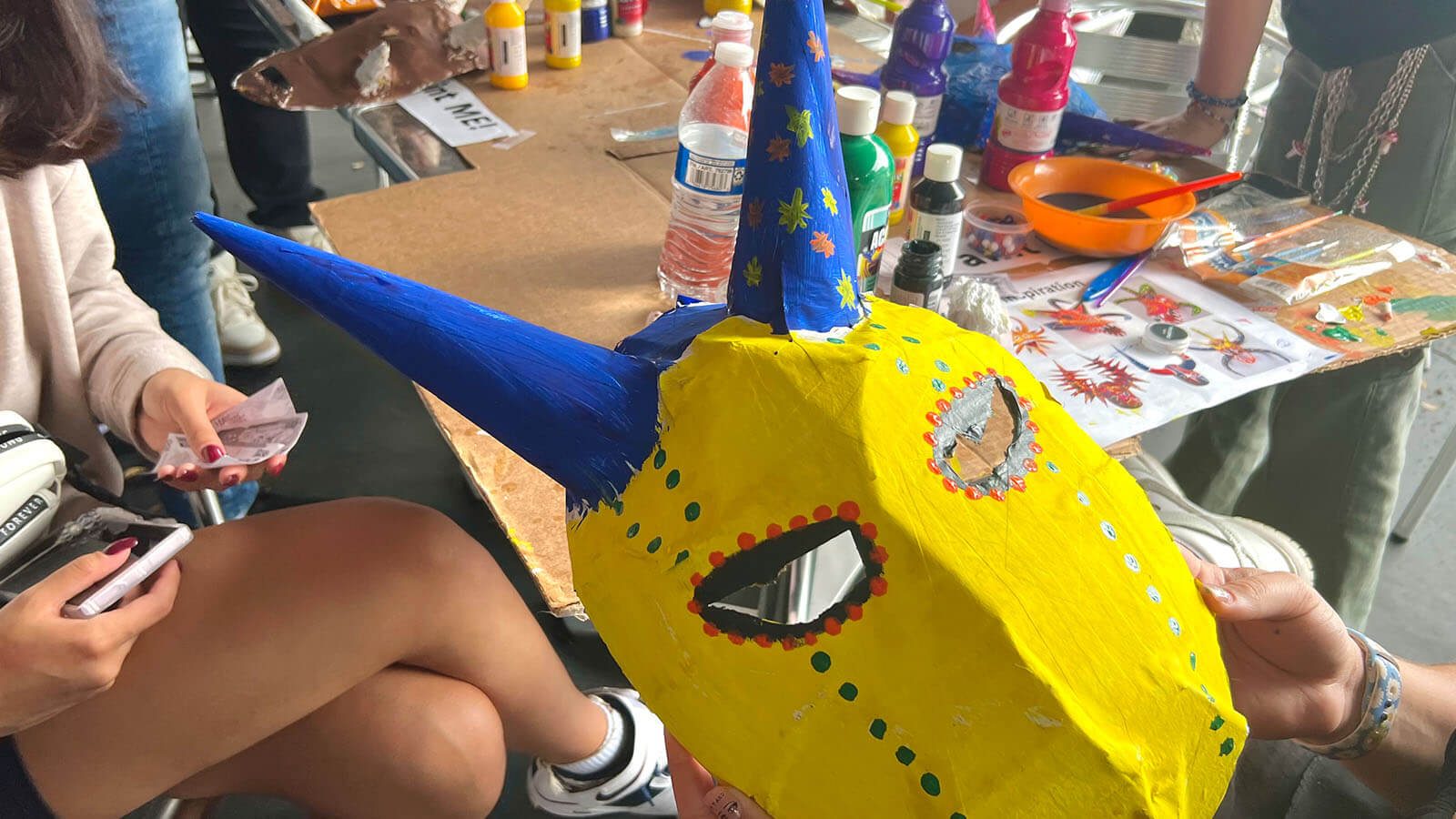 A person holds up a hand painted horned mask in front of a crowded arts and crafts table.