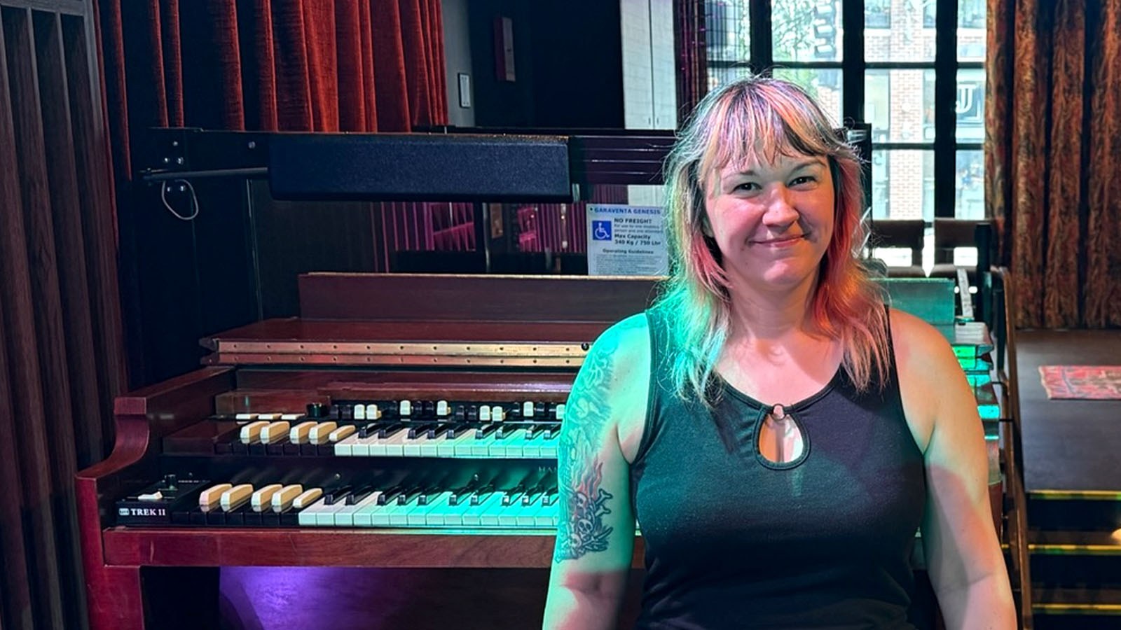 Kim Kozak sits on a piano bench in a backstage area at Garcia’s. She is smiling and wearing a black dress and tights.
