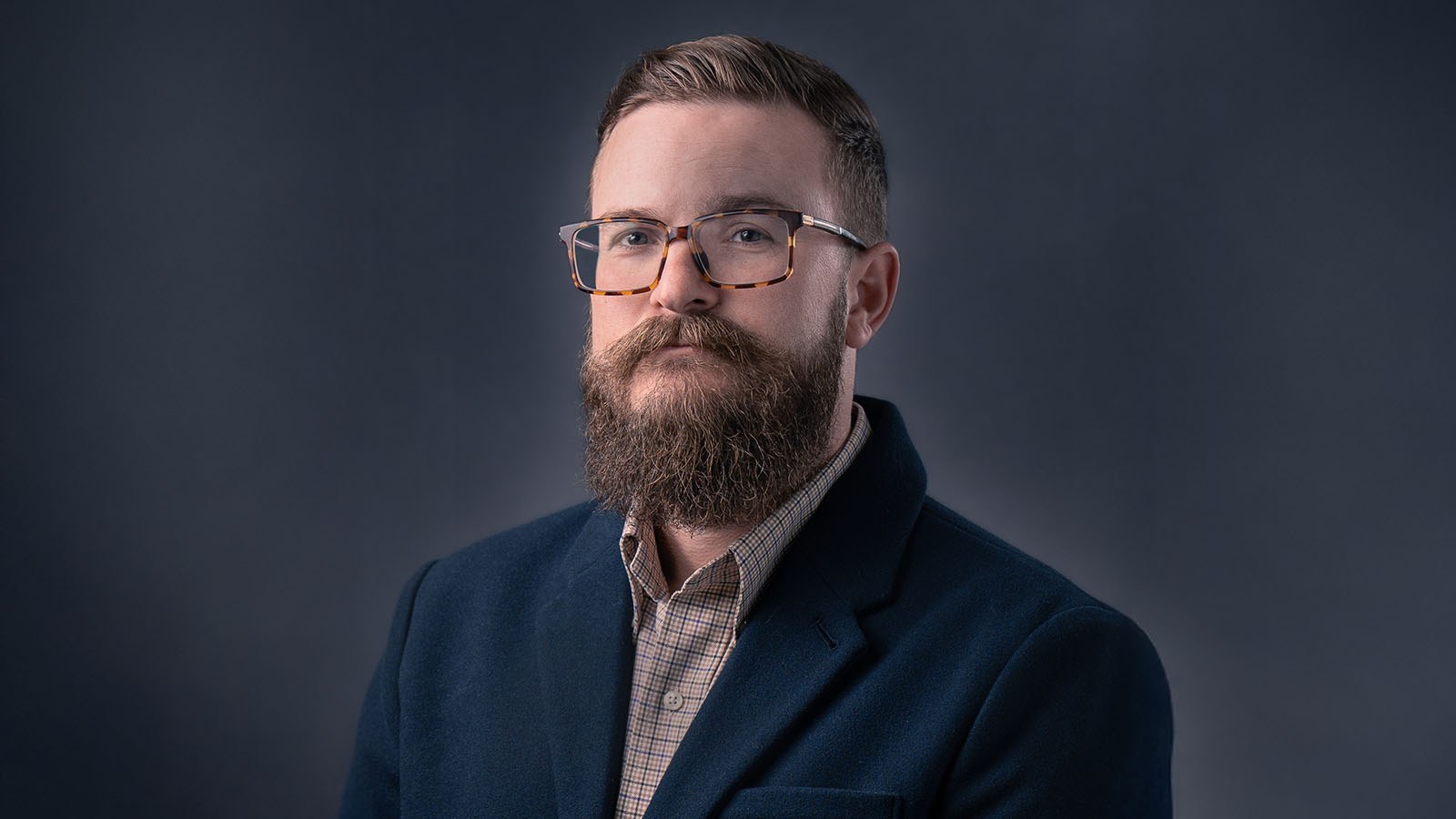 Drew Bailey wears a blue blazer and a brown checked shirt. He has brown hair, a beard, and glasses, and he’s smiling at the camera.