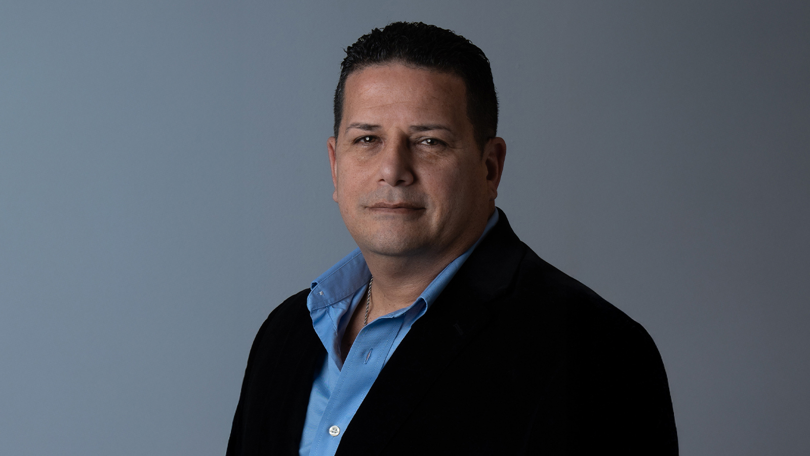 Sandy Navarro smiles at the camera in front of a grey background. He is wearing a blue button-down shirt and a black blazer.