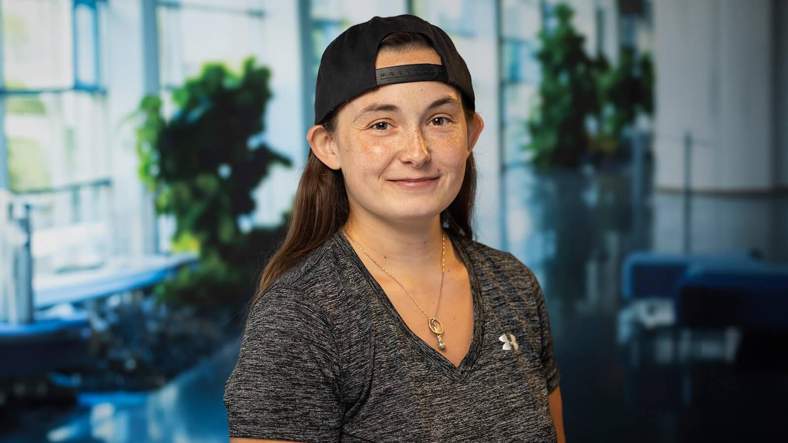 A person wearing a backward cap and a gray Under Armour shirt smiles in a bright, modern indoor space with plants in the background.
