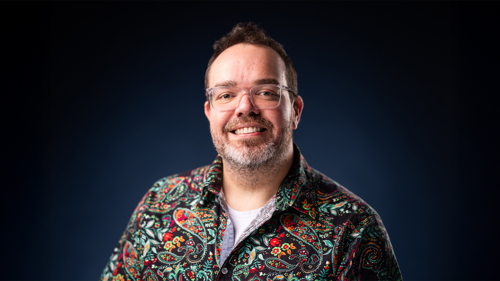 Tom Boldman smiles at the camera in front of a dark background. He is wearing a colorful paisley shirt and glasses.
