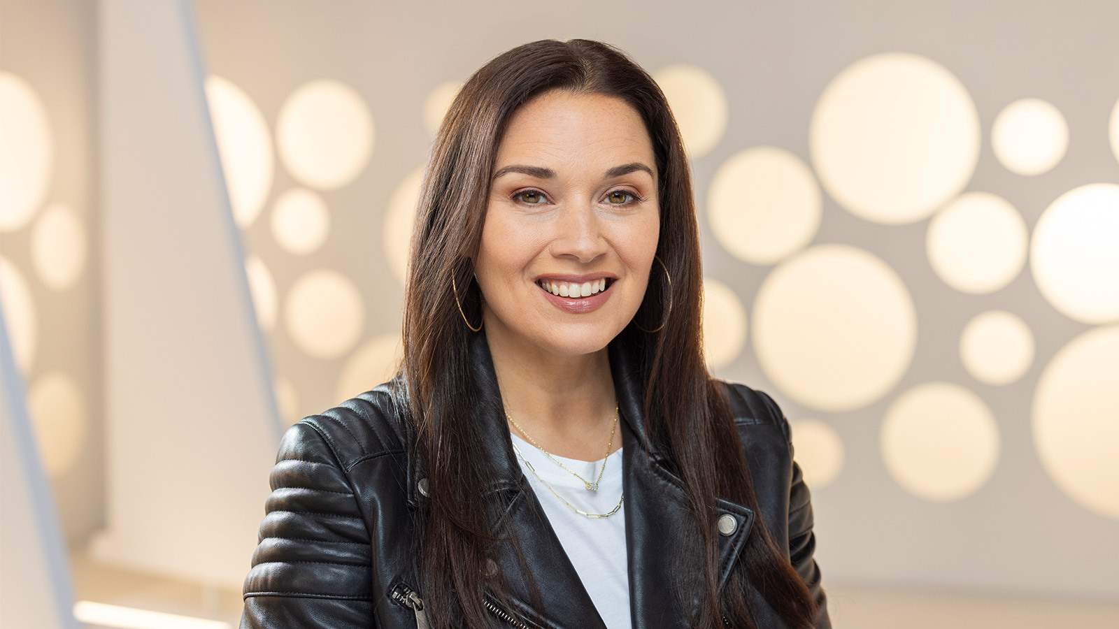 A headshot of a woman with straight brown hair and hoop earrings smiling while wearing a leather motorcycle jacket against a white background.