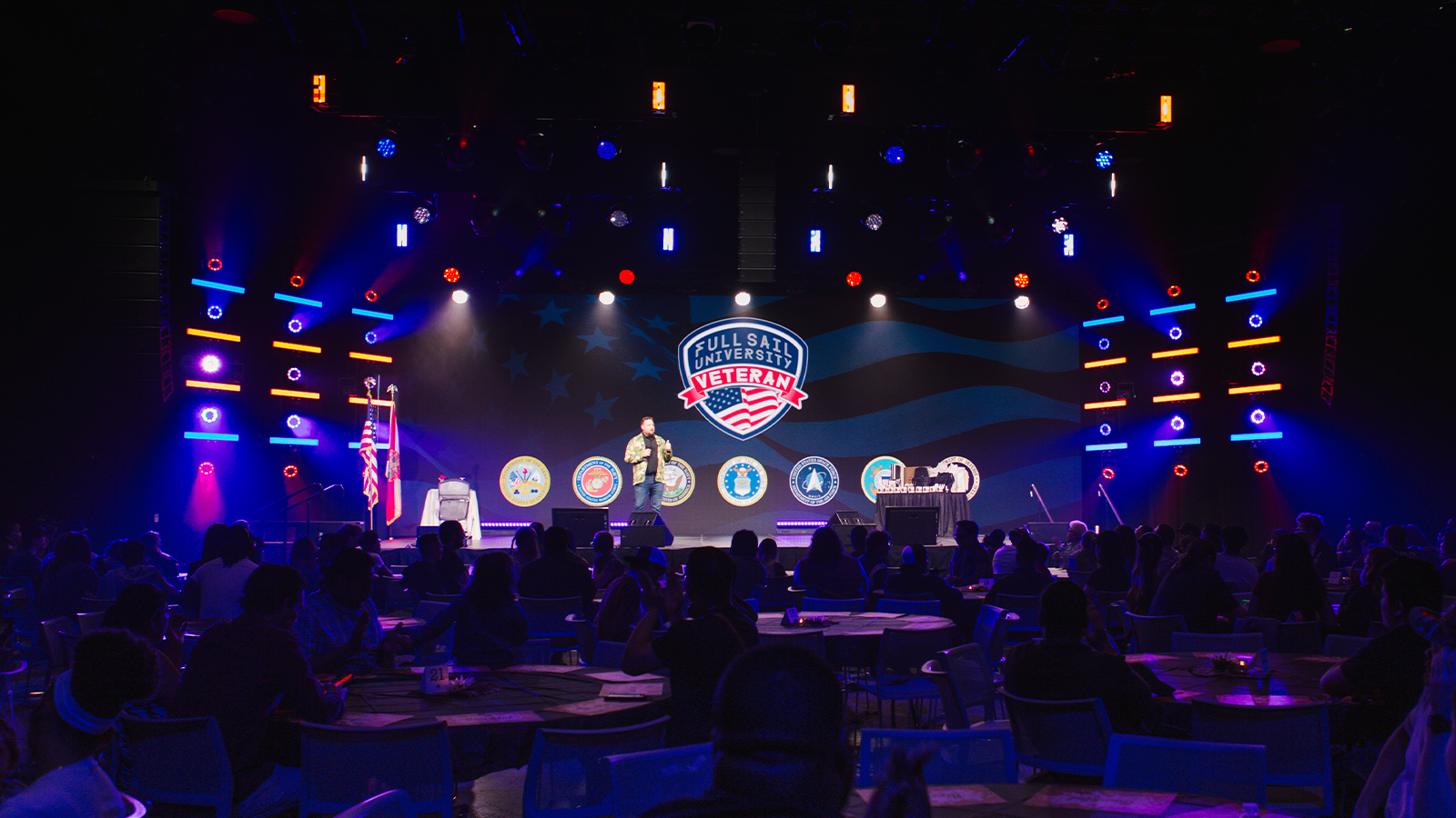 A crowd of people sit at round tables in a large room. A speaker stands on a stage holding a microphone with a screen behind them displaying the Full Sail University Veteran logo and logos of the branches of the military.