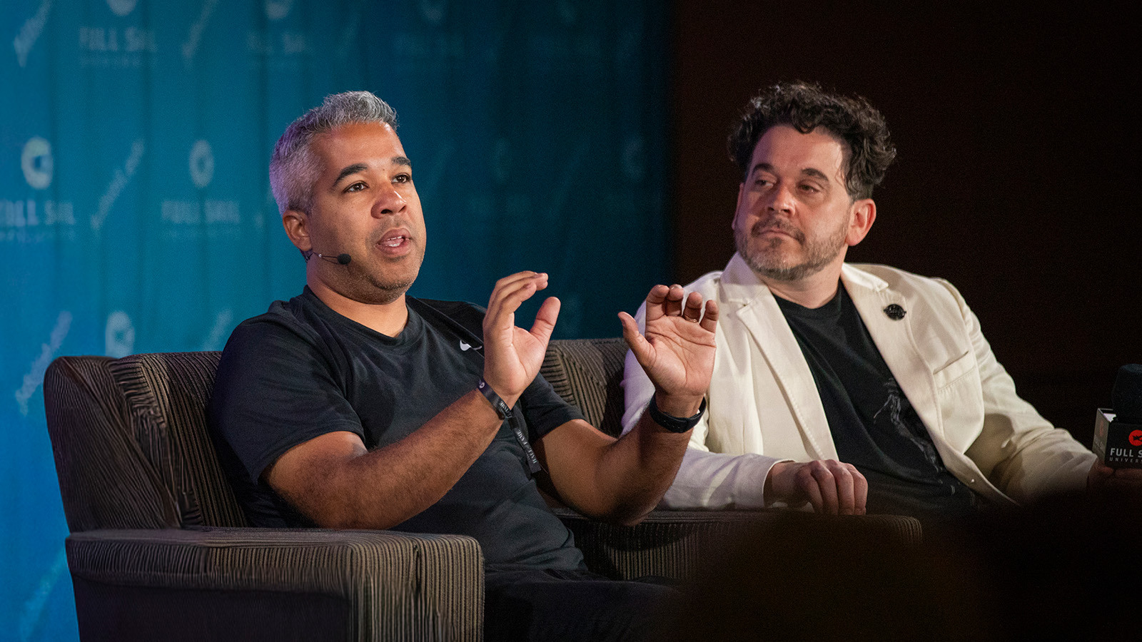 Juan Peralta and Gary Rizzo sit in armchairs on a stage and give a presentation. Juan wears a black t-shirt and Gary wears a black shirt and white blazer.