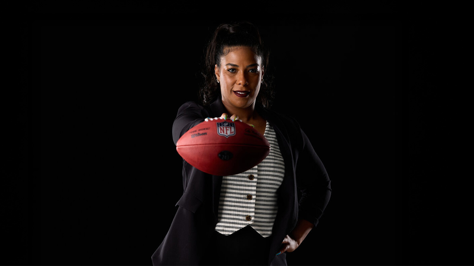 A woman in a blazer holds an NFL football toward the camera, confidently posing against a dark backdrop.