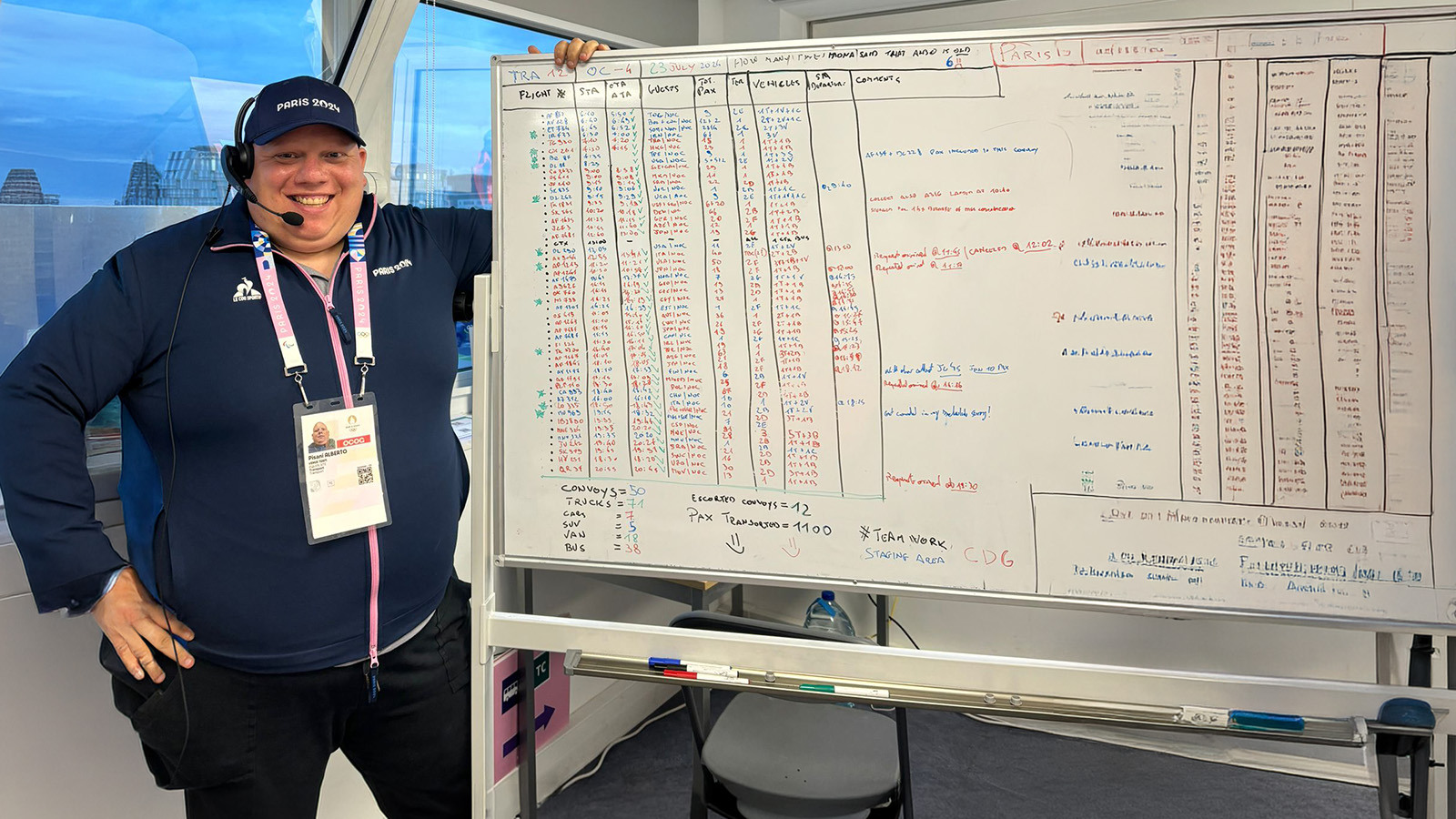 Alberto Pisani stands next to a large whiteboard with flight numbers on it and smiles. He wears a headset and a baseball cap that says Paris 2024.