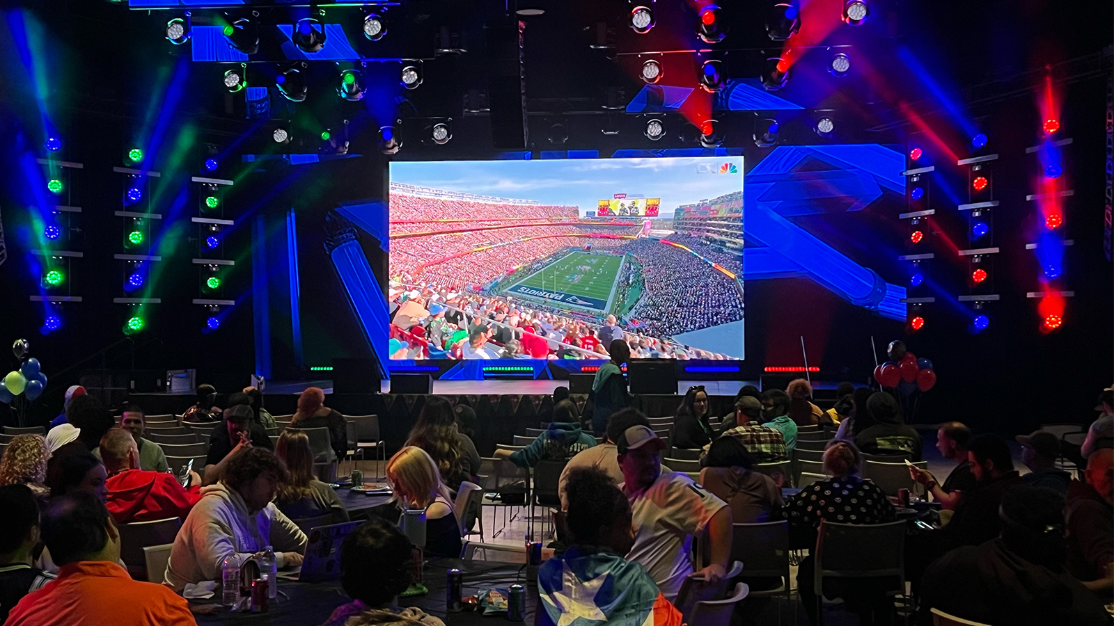 A crowd of students sit at round tables in a large room facing a large screen displaying a football stadium. One student wears a Puerto Rican flag as a cape.