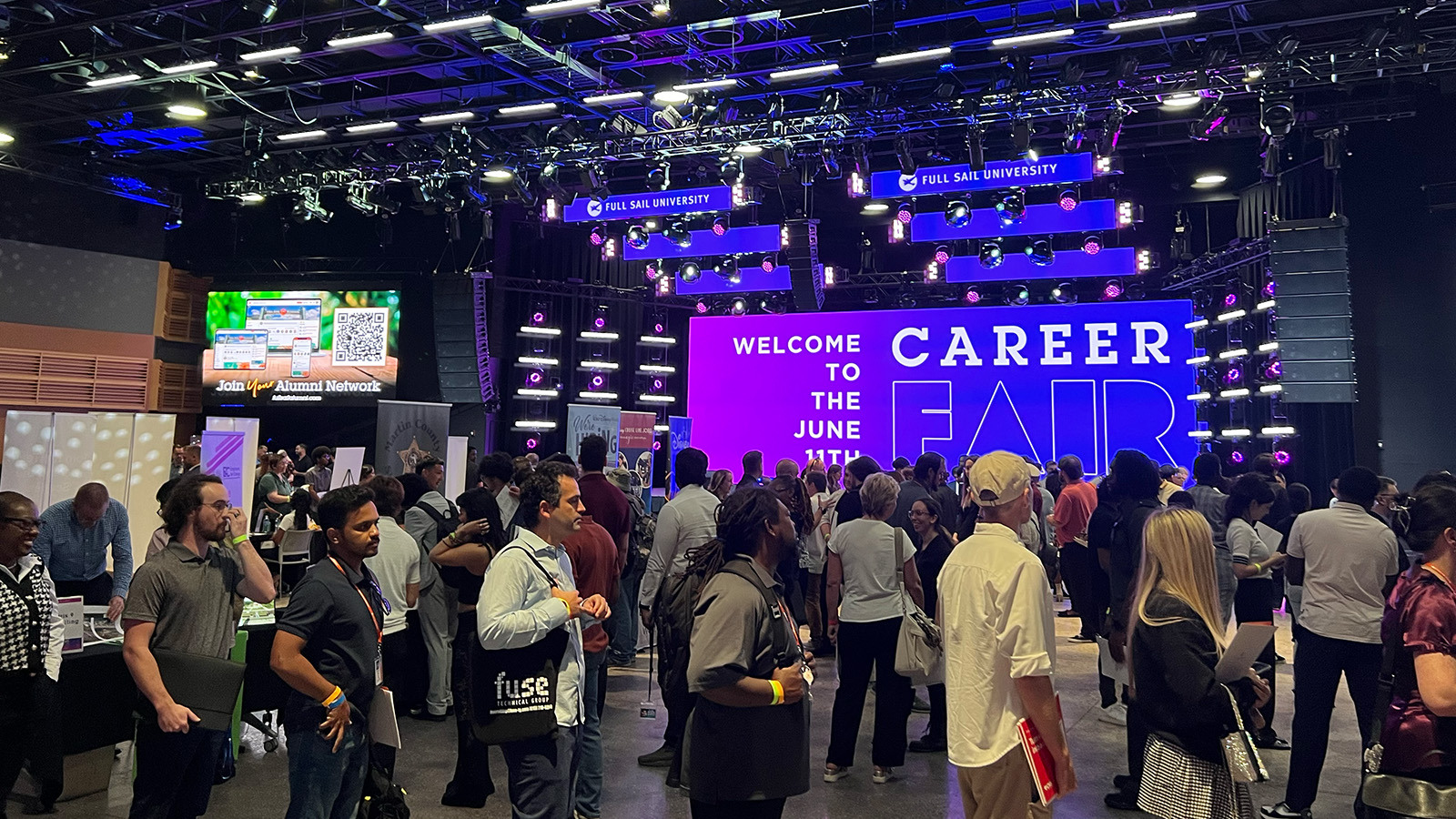 Students and alumni stand around and talk to potential employers. A screen behind them displays, “Welcome to the June 11th Career Fair.”