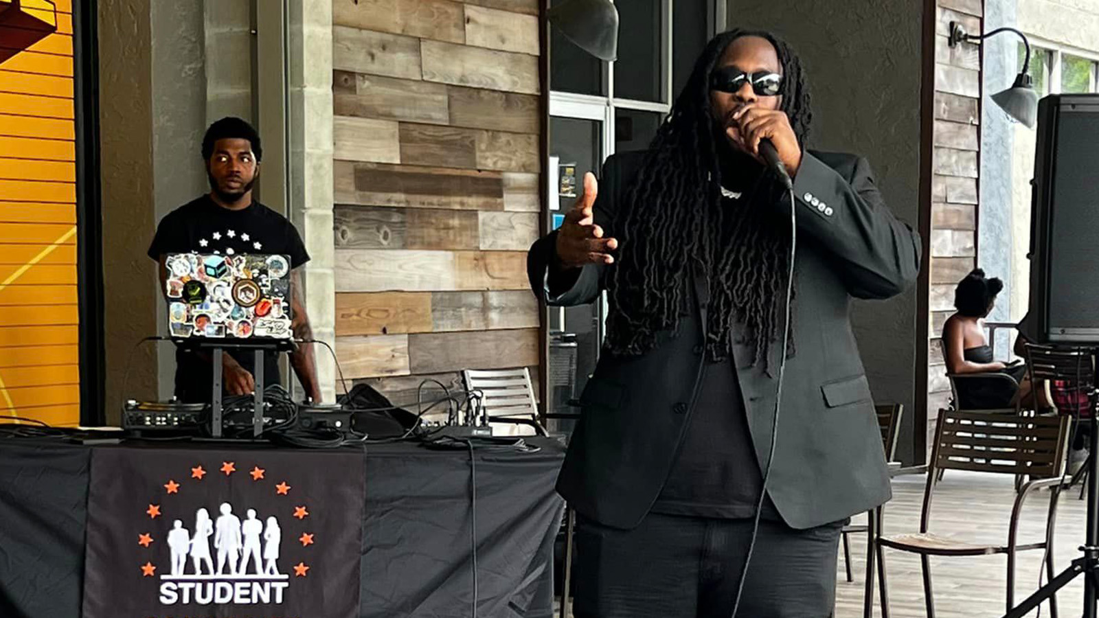 Guest speaker Souljah Bless, dressed in an all black outfit, speaks into a microphone on a patio stage. Behind him, a DJ stands at a booth that reads “Student Community Association.