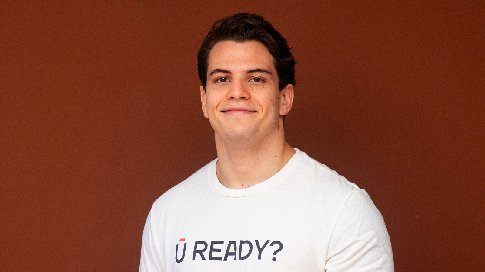 Gabriel Garnier smiles at the camera. He is wearing a white t-shirt that says “U READY?” and standing against a brown backdrop.