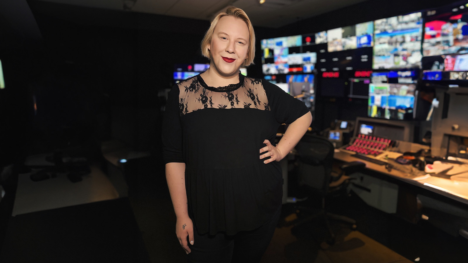 A person with blonde hair and a black lace-accent top stands in a dim control room, smiling, with colorful broadcast screens glowing behind them.