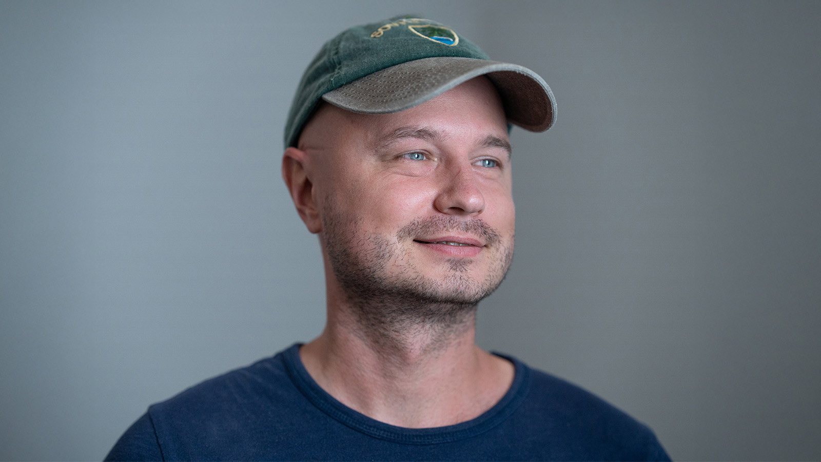 A portrait of a man wearing a green baseball cap and navy shirt while looking off to the side against a grey-green backdrop.