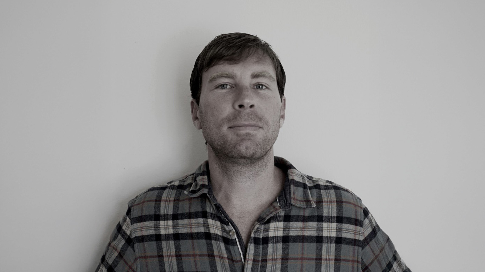 A man in a plaid shirt stands against a plain wall, looking directly at the camera with a very slight smile.