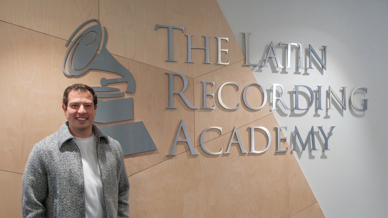 Nahun Paguada stands in front of a wall with a large sign displaying “The Latin Recording Academy” with a gramophone logo. He is wearing a white shirt, grey jacket, and khaki pants with his hands in his pockets.