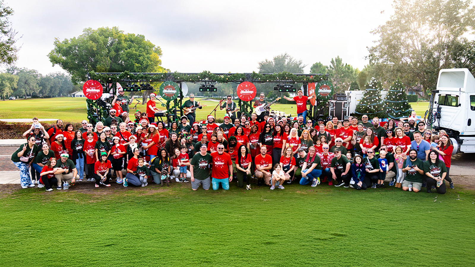 A large group of staff, faculty, and their families pose for a photo wearing red and green holiday shirts in front of a semi-truck bed decorated with garlands and lights. Some stand on the truck bed holding various musical instruments.