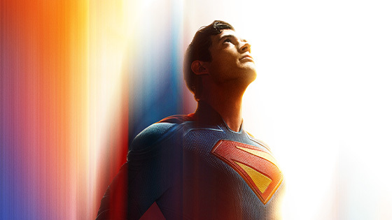 Superman stands and looks at the sky. He is wearing his blue costume with a yellow and red S on the front and his red cape is billowing behind him.