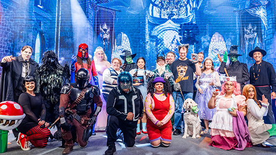 A group of staff members pose for the camera dressed in Halloween costumes including Glinda and Elphaba, the Mandalorian, Loonette the Clown, and more.