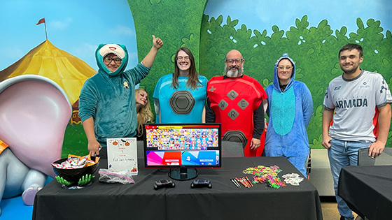 Full Sail staff and students stand next to each other and smile at the camera wearing costumes. A table in front of them has candy, stickers, and a gaming console on it.