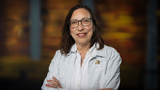 Abby Ginsburg smiles at the camera in front of a wood background. She is wearing a white jacket and glasses.