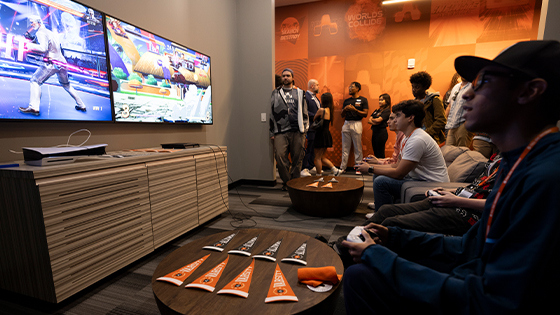 Gamers compete in a lounge with dual screens, a PlayStation 5, and Armada-branded decor, while a crowd watches in the background.
