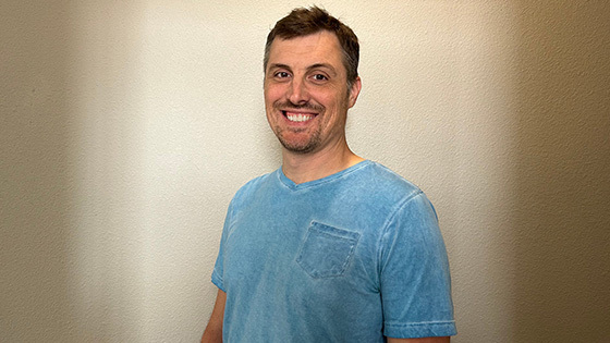 James Lane stands in front of a beige wall and smiles at the camera. He is wearing a light blue v-neck t-shirt.