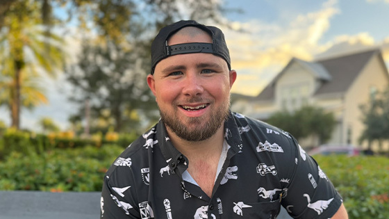 Tyler Yates sits on a bench in an outdoor park and smiles. He’s wearing a black and white collared shirt with dinosaurs on it and a backwards cap.