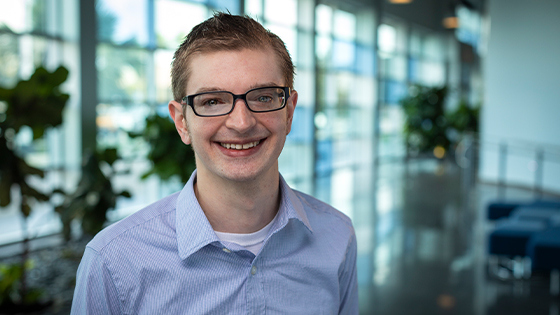 Wes Locher stands in a lobby with lots of windows and smiles at the camera. He wears glasses and a blue checked button-up shirt.