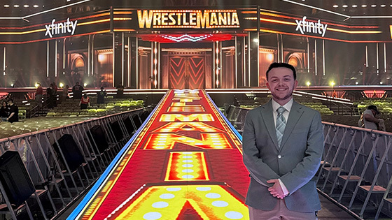 Colby Applegate wears a suit and stands in a brightly lit wrestling arena with Wrestlemania graphics displayed on the walls and floor.