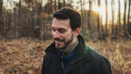 Mikey Zoppo stands in a forest with sunlight poking through the trees behind him. He looks down and smiles, and he is wearing a dark jacket.