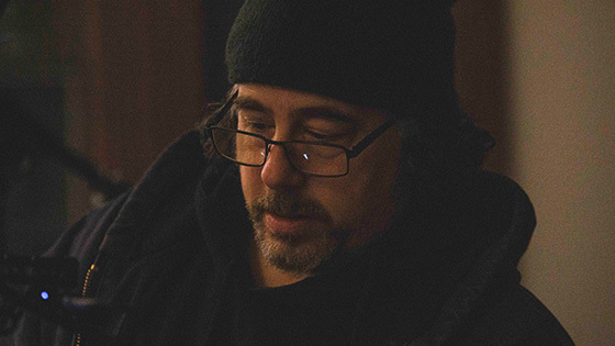Michael Romano stands in front of a grey wall wearing a beanie, hoodie, and glasses with a pencil tucked behind his ear.