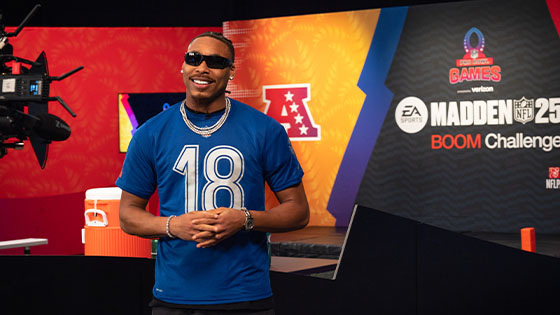 Minnesota Vikings wide receiver Justin Jefferson smiles while wearing a blue NFL-branded t-shirt with the number '18' in a studio set for the Madden NFL 25 Boom Challenge.