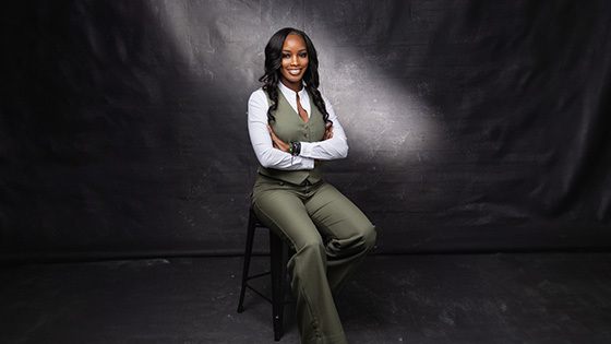 A woman in a green vest and pants sits confidently on a stool, arms crossed, against a dark studio backdrop.