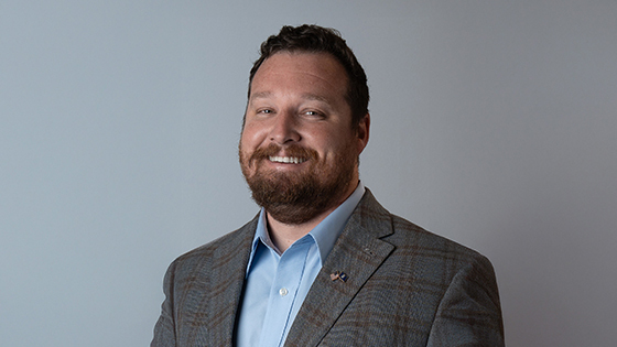 A man with short brown hair and a short brown beard smiles at the camera, wearing a plaid blazer and light blue shirt against a grey background.