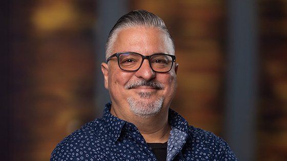 Alex Ginzo smiles at the camera. He is wearing a blue and white collared shirt and glasses, and he has gray hair and a short gray beard.