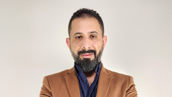 Dr. Ahmed Al Zaidy stands with his arms crossed and smiles at the camera. He is wearing a brown blazer over a blue button-up shirt.