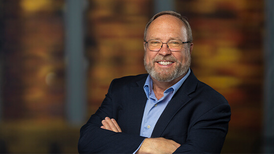 Reo McBride stands with his arms crossed and smiles at the camera. He has a beard and glasses; he’s wearing a blue button-up shirt and a blue blazer.