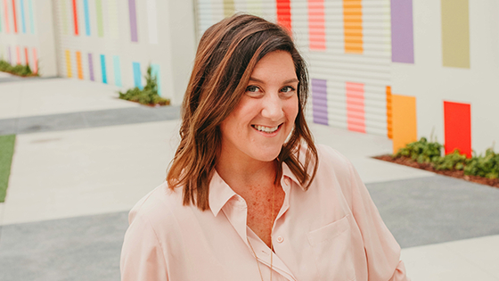 Jenn Miller sits in front of a colorful outdoor wall. She is smiling and wearing a pink shirt and a gold necklace.