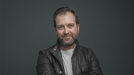 A man in a dark brown leather coat and light t-shirt appears from the waist up, smiling against a grey backdrop.