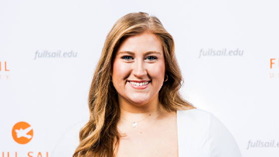 A woman smiles while holding a Full Sail 5 year anniversary guitar pick in front of a Full Sail University step-and-repeat backdrop.
