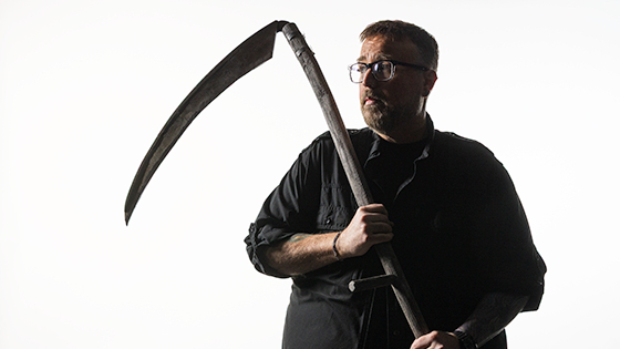 Full Sail Grad Aaron B. Koontz is seen wearing black as he holds a large scythe against a bright white background, gazing thoughtfully to the left side of the frame.