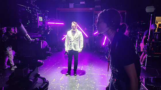 An actor stands in the middle of a set wearing a white suit and bowtie with purple lights around them. A student wears a headset and operates a camera pointed at the actor.