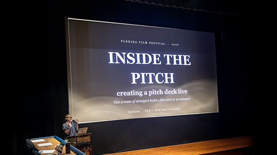 Anne Russell speaks at a podium in front of a large screen displaying “Florida Film Festival 2026 Inside the Pitch: creating a pitch deck live. Can a room of strangers build a film pitch in 30 minutes?”