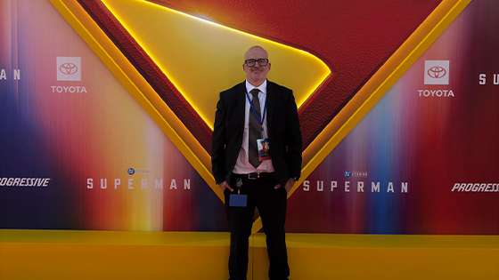 Grad Guillermo Cummings stands before a glowing Superman logo backdrop at a movie premiere, wearing a lanyard and event badge.