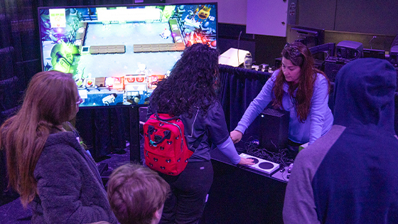 Several people stand around facing a table with adaptive gaming consoles, speakers, and a large screen nearby displaying a game. A staff member stands behind the table.