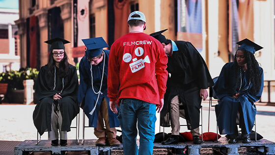A Coca-Cola Refreshing Films crew member faces seated graduates in caps and gowns during a staged production scene.