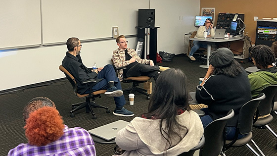 Brad Biwer sits in a chair and speaks to a crowd of students. A staff member sits in a chair next to him and a faculty member sits at their desk behind them.