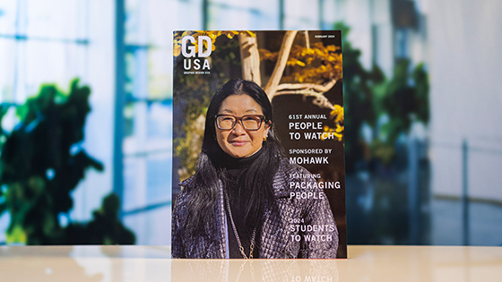 A copy of “Graphic Design USA” magazine depicting a woman wearing glasses and a jacket stands atop a white table.