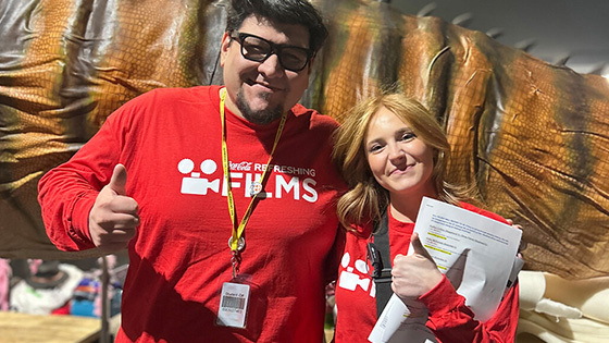 Director Angel and Producer Kylie smile in front of a large animatronic dinosaur tail on set during their film shoot.